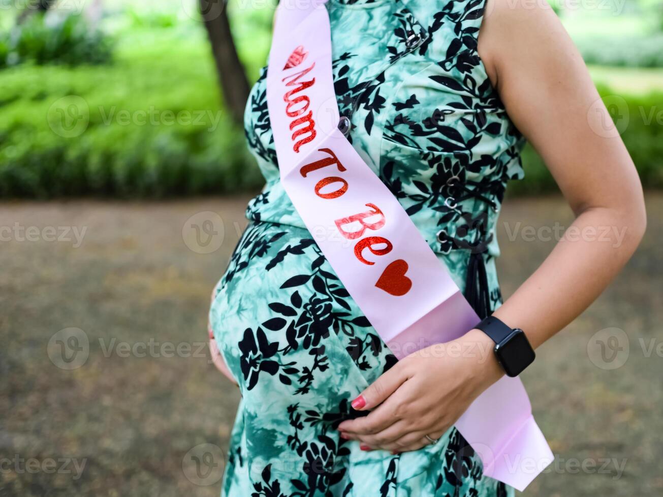 uma grávida indiano senhora poses para ao ar livre gravidez tiro e mãos em barriga, indiano grávida mulher coloca dela mão em dela estômago com uma maternidade vestir às sociedade parque, grávida lado de fora maternidade tiro foto