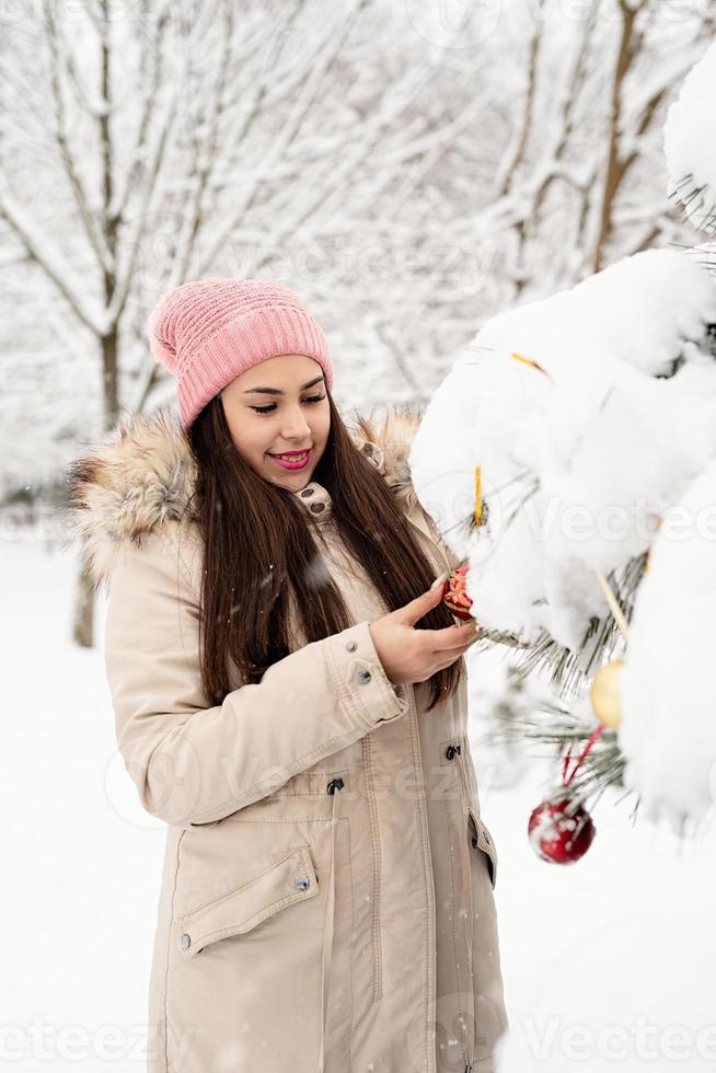 Mulher bonita em roupas quentes de inverno decorando uma árvore de Natal em um parque em um dia de neve foto