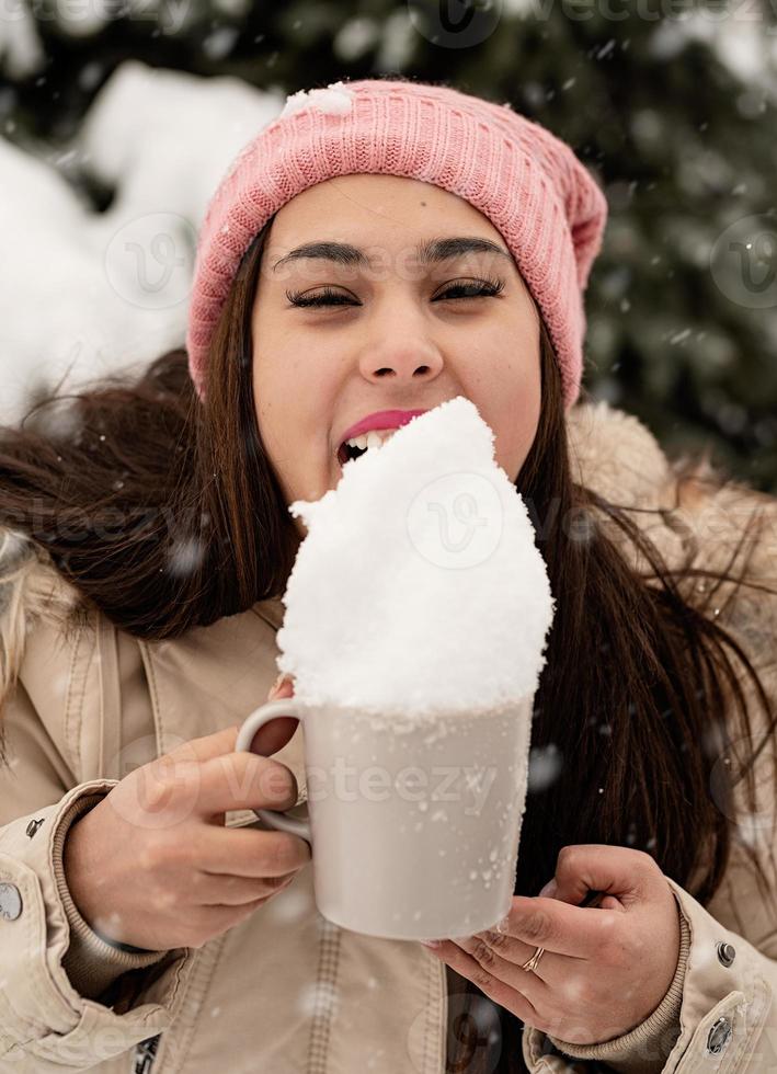 mulher com roupas quentes de inverno em pé ao lado da grande árvore de Natal ao ar livre e lambendo a neve da xícara foto