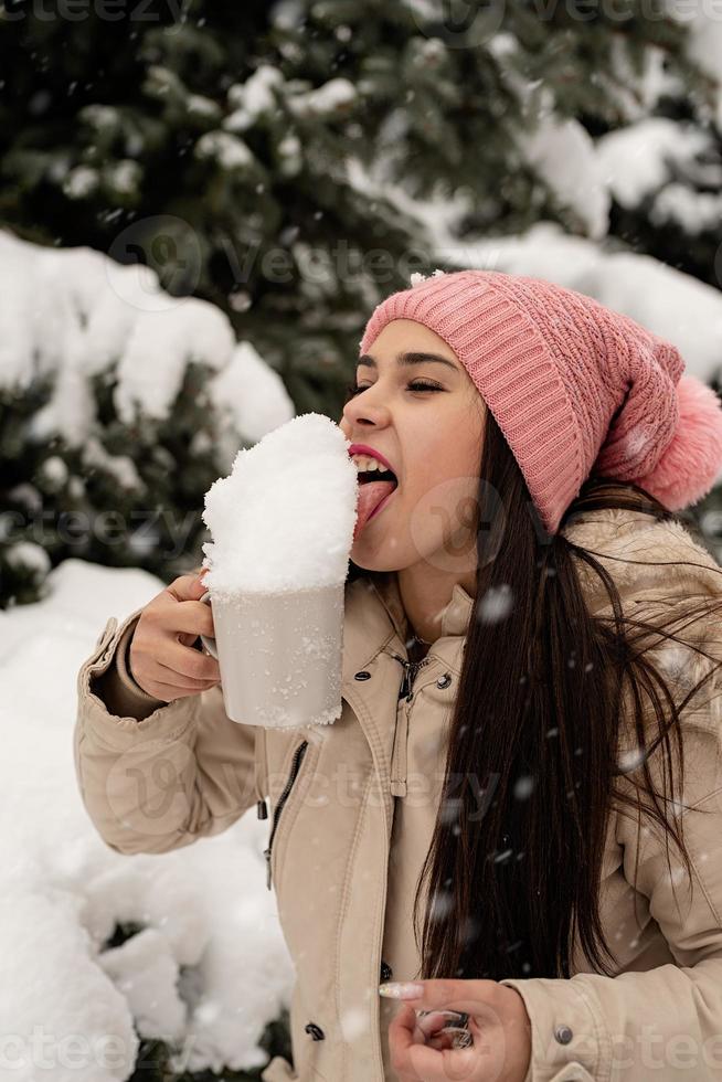 mulher com roupas quentes de inverno em pé ao lado da grande árvore de Natal ao ar livre e lambendo a neve da xícara foto