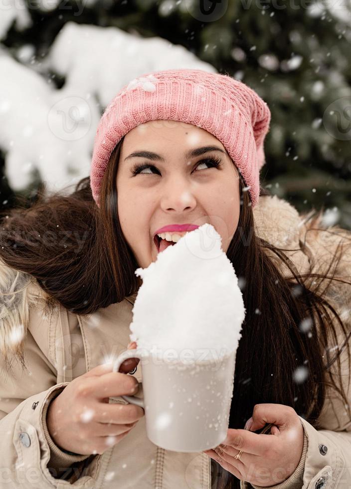 mulher com roupas quentes de inverno em pé ao lado da grande árvore de Natal ao ar livre e lambendo a neve da xícara foto