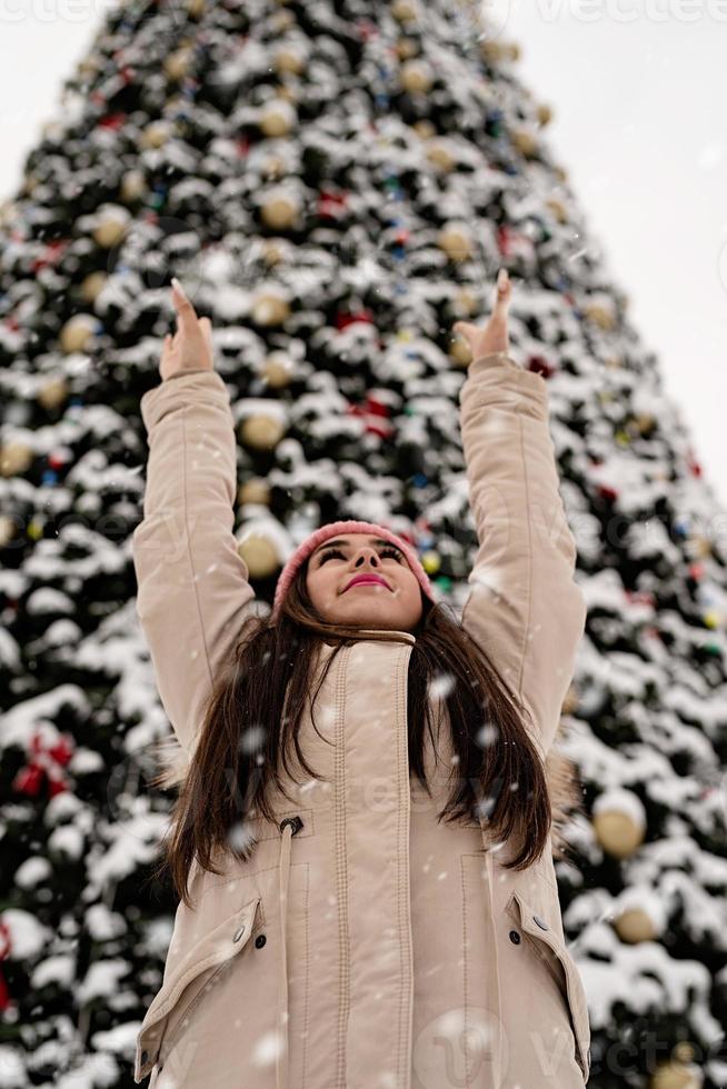 mulher com roupas quentes de inverno em pé ao lado da grande árvore de Natal ao ar livre, braços para cima, neve caindo foto