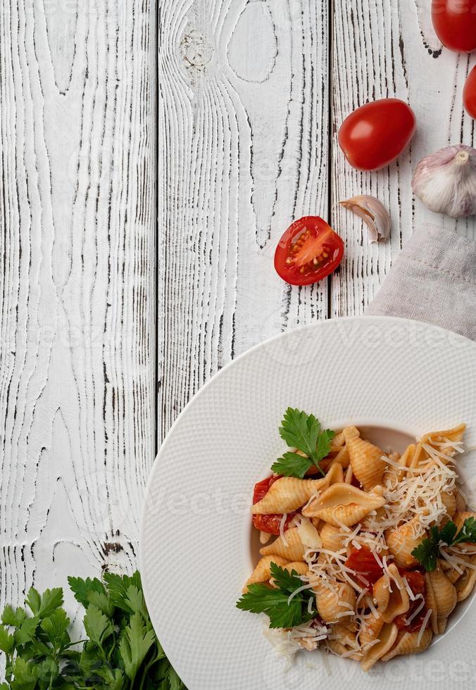 Macarrão conchiglioni com molho à bolonhesa com tomate cereja, queijo e salsa em prato branco sobre fundo de madeira foto