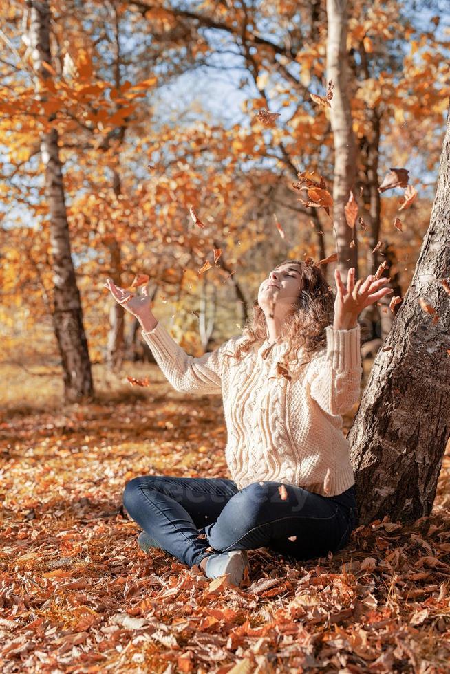 jovem jogando folhas amarelas para cima, sentada na floresta de outono foto