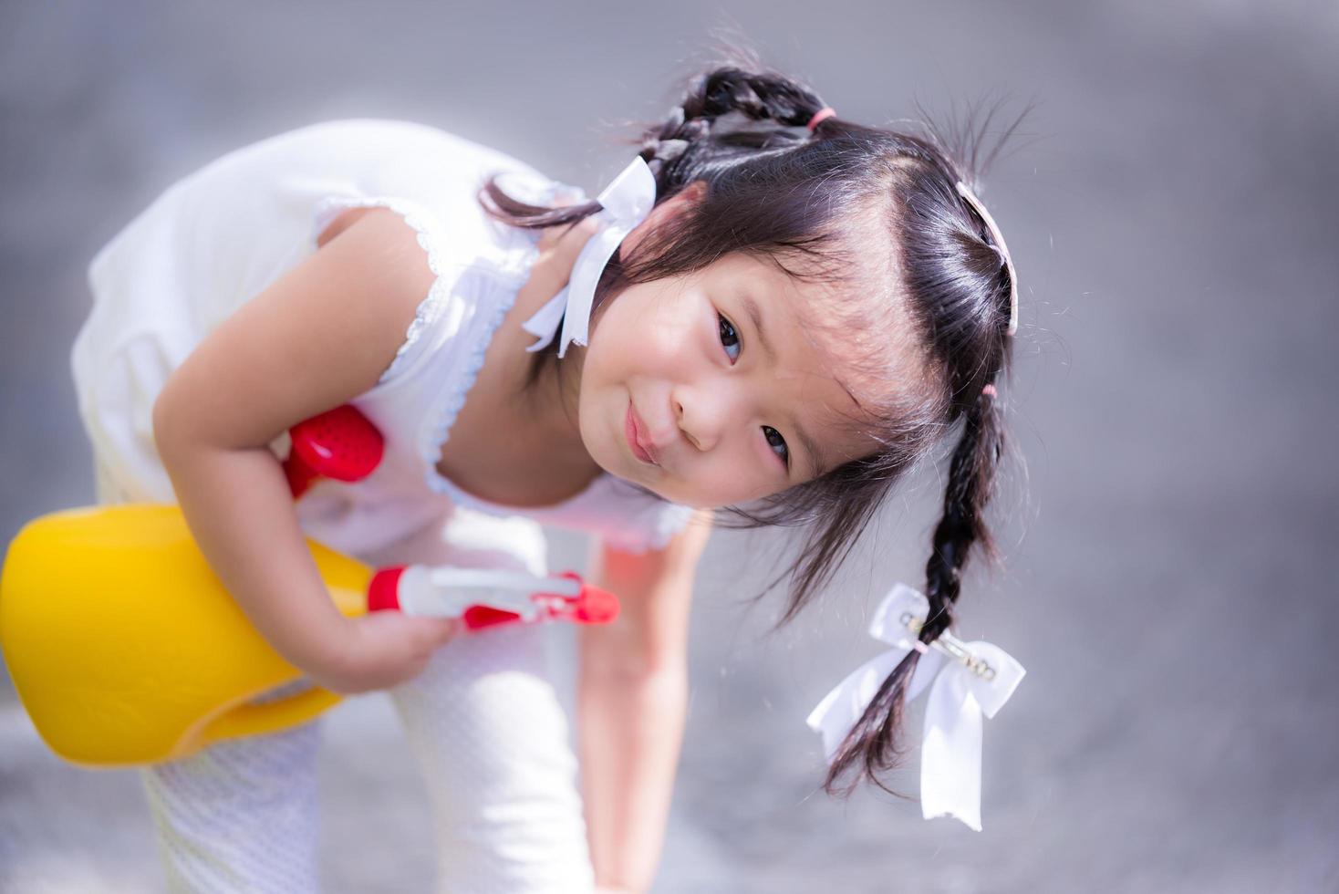 linda garota brincando com um regador de planta amarela. criança sorriu docemente e coçou as pernas. garotinho vestido de branco, 3 anos. foto