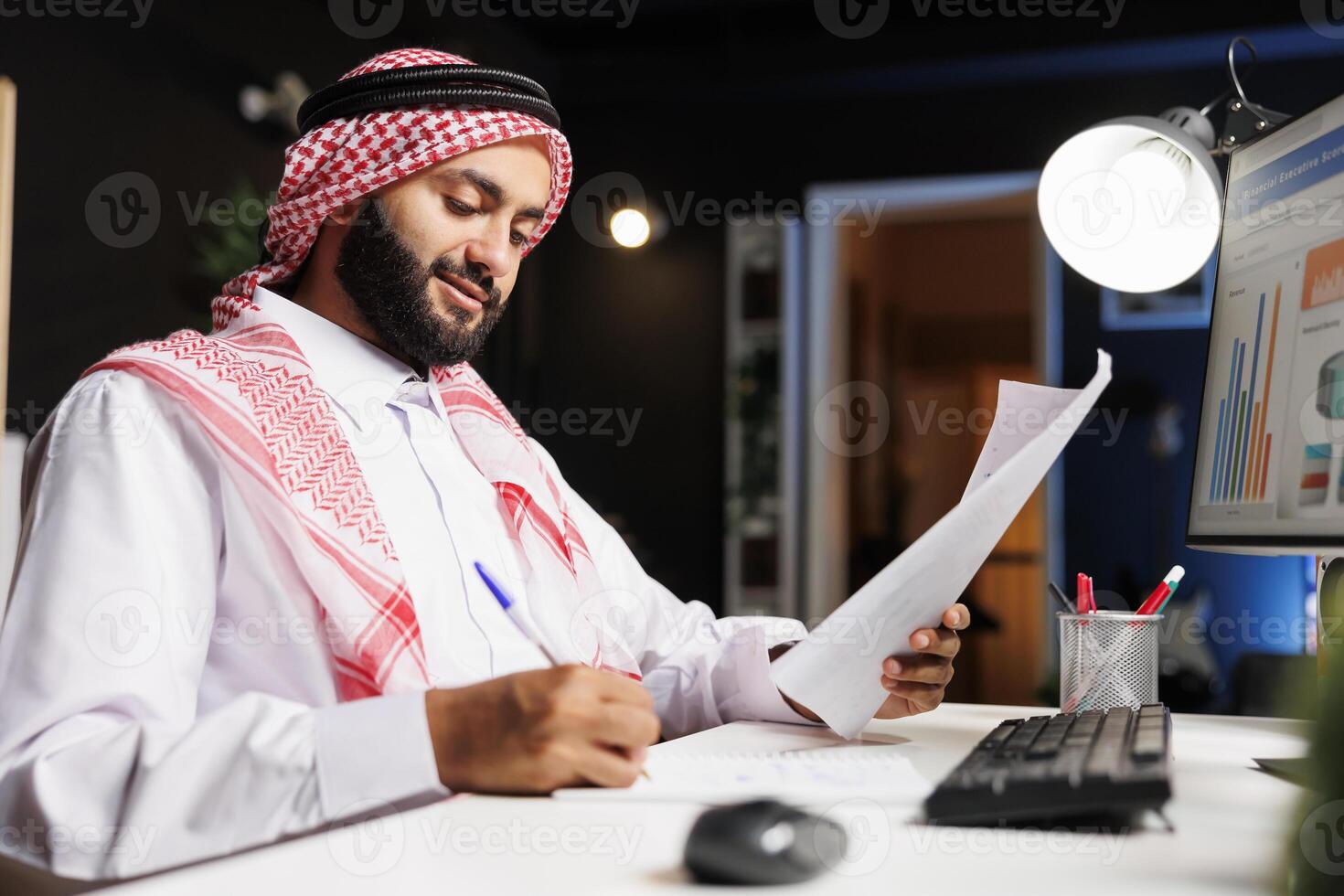 islâmico homem trabalhando às dele mesa, cercado de uma Área de Trabalho computador, documentos, e papelada, exibindo uma ocupado corporativo área de trabalho. vista lateral tiro do muçulmano cara escrevendo baixa notas a partir de pesquisar. foto