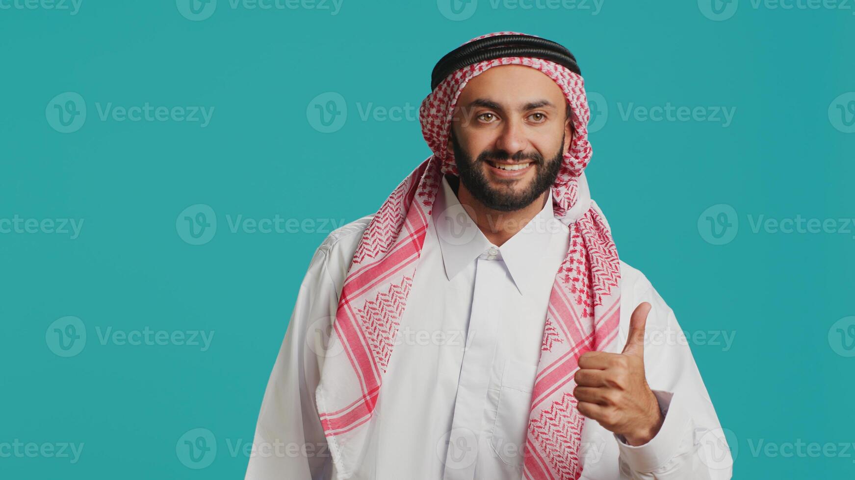 confiante homem mostrando gostar placa dentro estúdio, dando polegares acima e acordo enquanto ele representa tradicional árabe cultura. meio Oriental positivo cara desgasta islâmico traje e lenço de cabeça. foto
