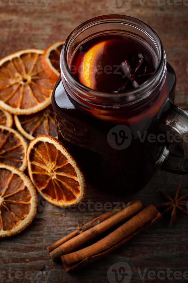 vinho quente caseiro, laranjas secas e paus de canela em uma mesa de madeira sobre um fundo escuro foto