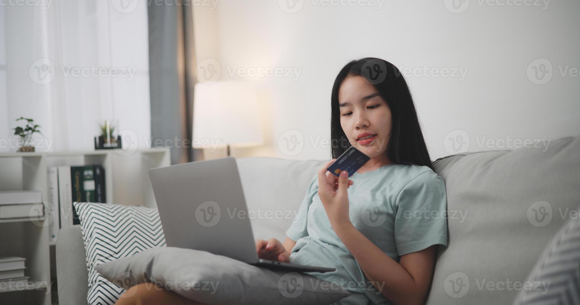seletivo foco, jovem ásia mulher sentado em sofá segurando crédito cartão fazer conectados Forma de pagamento para compra dentro rede loja usando computador portátil. foto