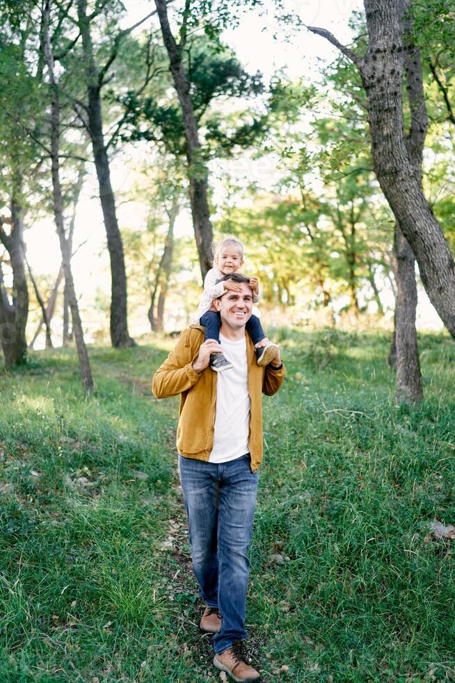 Papai com uma pequeno menina em dele ombros anda em através a floresta foto