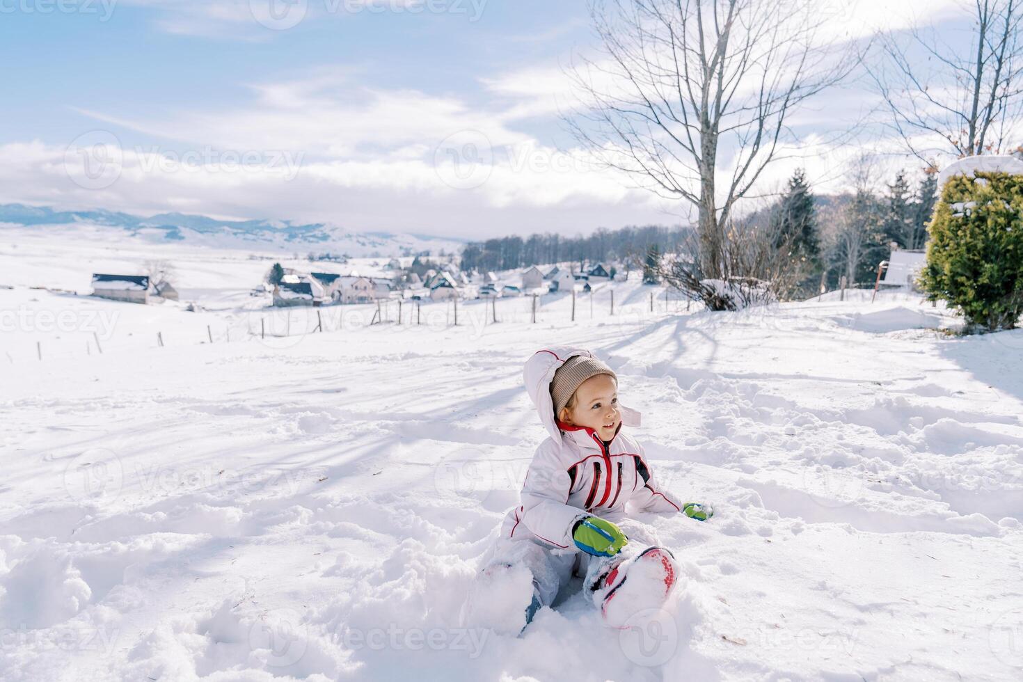 pequeno menina senta meio virado dentro uma monte de neve e parece longe foto