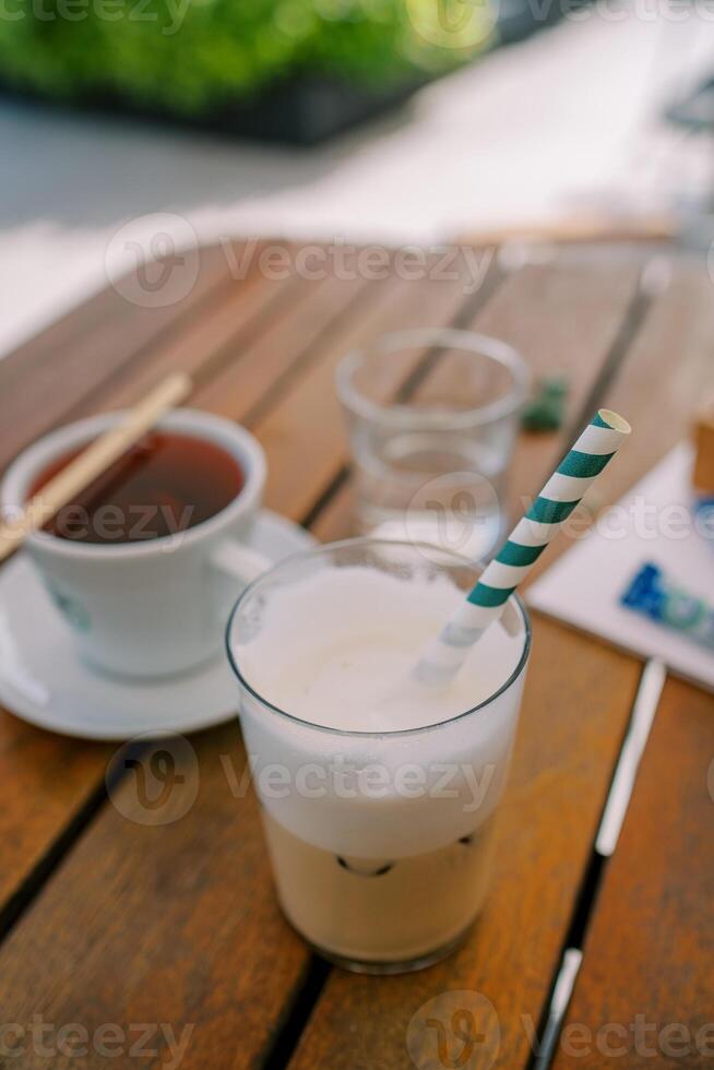 vidro do cappuccino com uma listrado Palha carrinhos em uma de madeira mesa Próximo para uma copo do chá foto