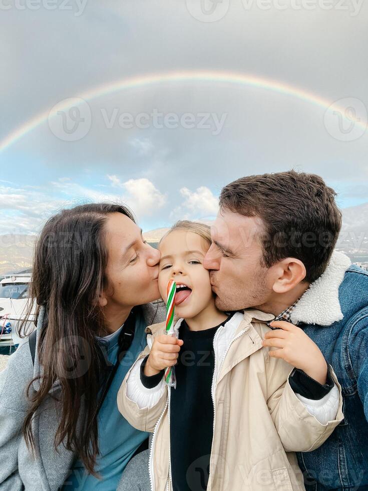 Papai e mãe beijo em a bochechas uma pequeno menina com uma pirulito em uma arco Iris fundo foto