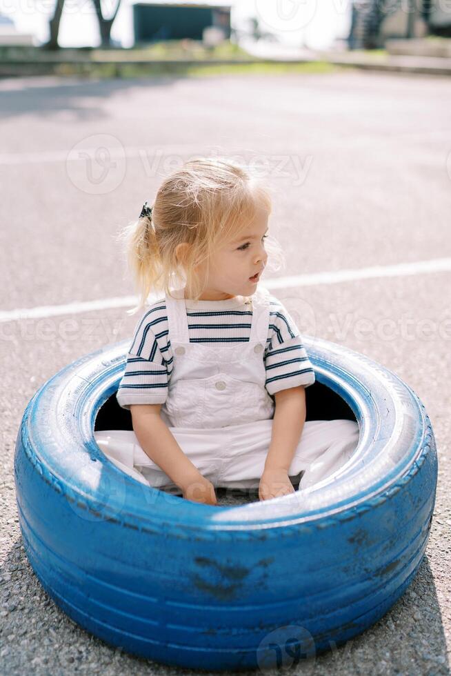 pequeno menina senta dentro uma grande azul pneu em a Parque infantil e parece longe foto