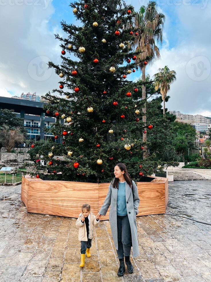 mãe com uma pequeno menina ficar de pé em a rua perto uma decorado Natal árvore foto