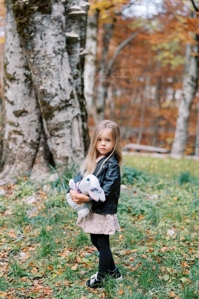 pequeno menina com uma brinquedo Coelho dentro dela mãos carrinhos meio virado dentro a outono floresta foto