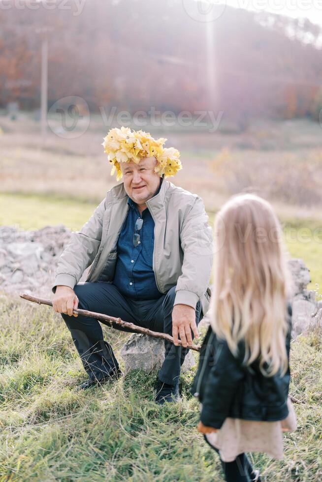 pequeno menina parece às dela avô sentado em uma pedra em a gramado dentro uma guirlanda do amarelo folhas. costas Visão foto