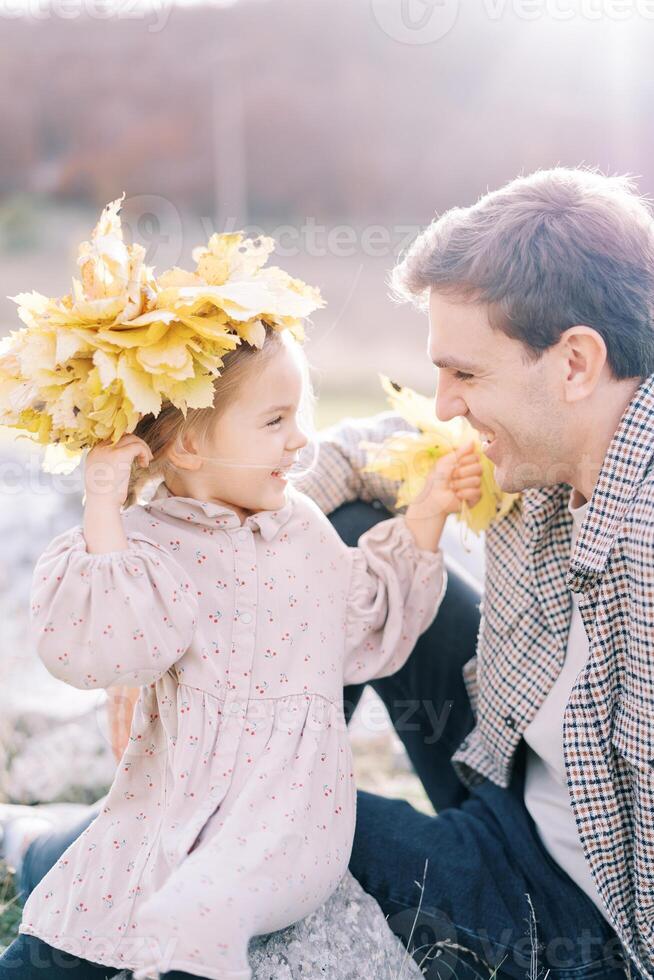 pequeno sorridente menina parece às dela Papai sentado em uma pedra dentro uma guirlanda do outono folhas foto