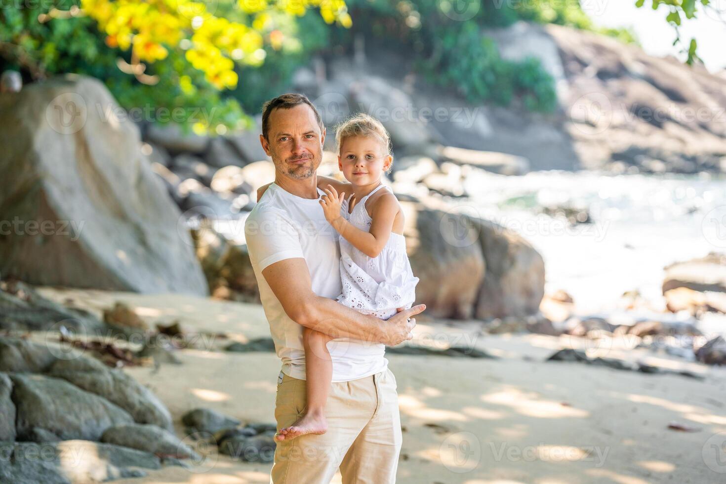 pequeno menina em do pai mãos sorrir e alegrar em Beira Mar dentro a sombra do árvores e Palmeiras. feliz família conceito. Alto qualidade foto