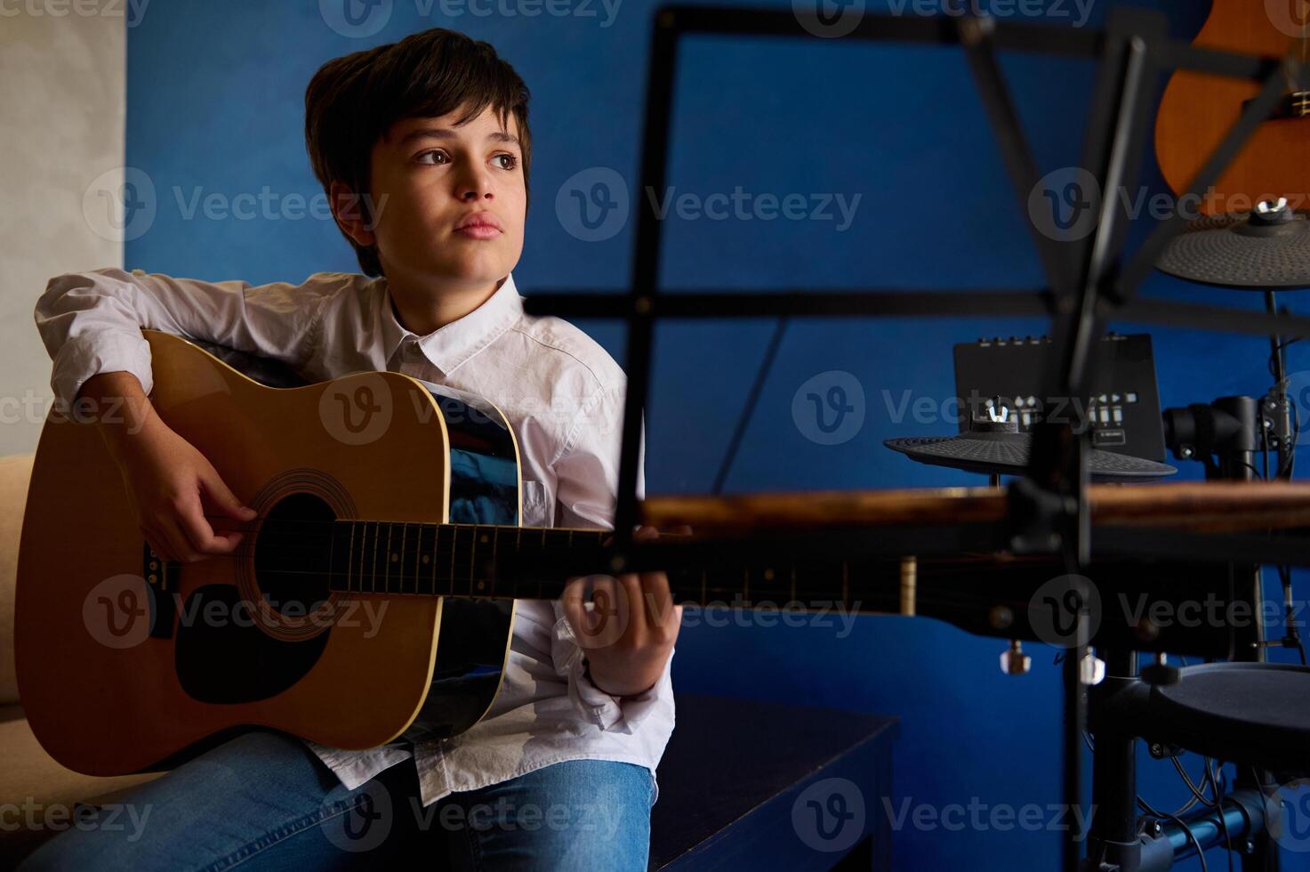 hispânico Adolescência Garoto dentro branco casual camisa, arrancar cordas enquanto dedilhar guitarra, jogando acústico clássico guitarra, sentado dentro a música estúdio sobre azul parede fundo e musical instrumentos foto