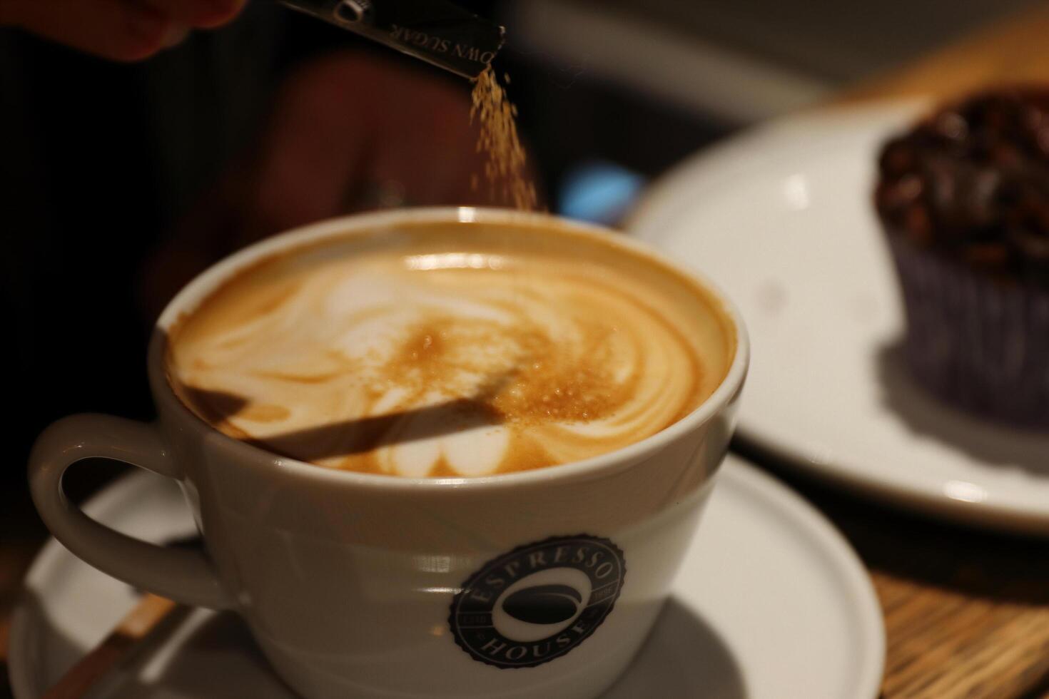 copenhague, Dinamarca - fevereiro, 20, 2023. espresso casa. adicionando açúcar para a copo do cappuccino. cremoso espuma e delicioso caseiro chocolate Bolinho em a vintage de madeira mesa. seletivo foco foto