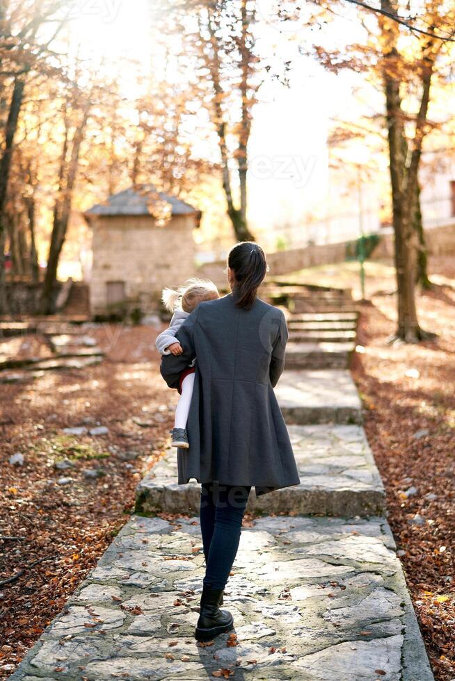 mãe com uma pequeno menina dentro dela braços anda em ao longo uma pavimentou caminho dentro a outono floresta. costas Visão foto