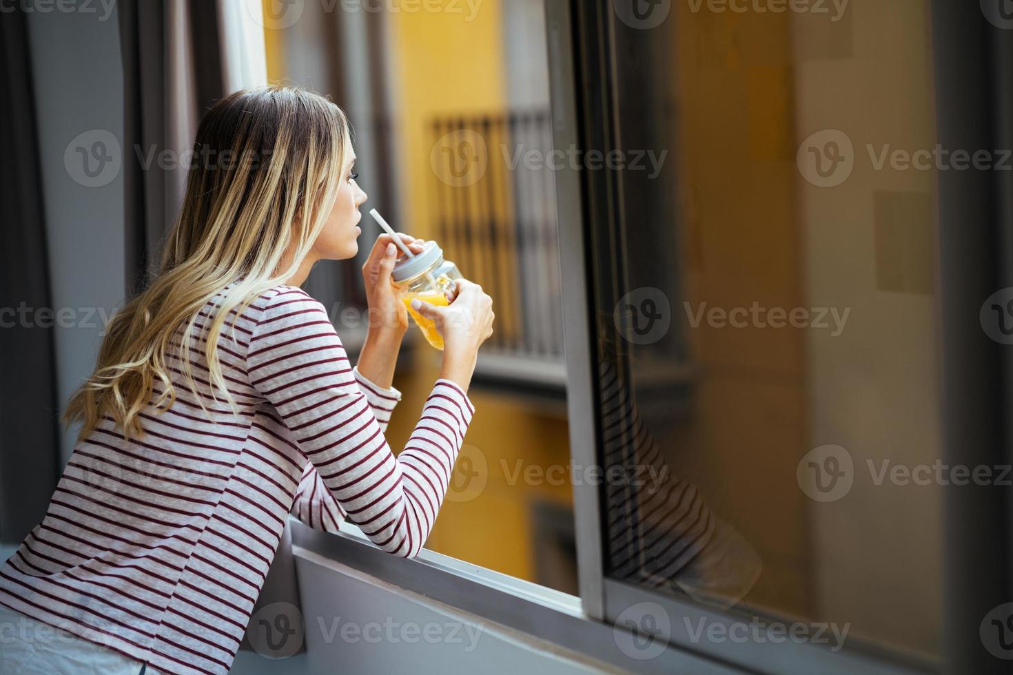 jovem bebendo um copo de suco de laranja natural, inclinando-se para fora da janela de sua casa. foto