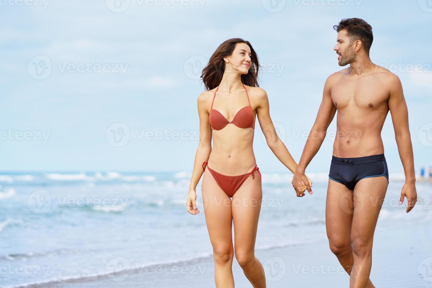 jovem casal de lindos corpos atléticos caminhando juntos de mãos dadas na praia foto