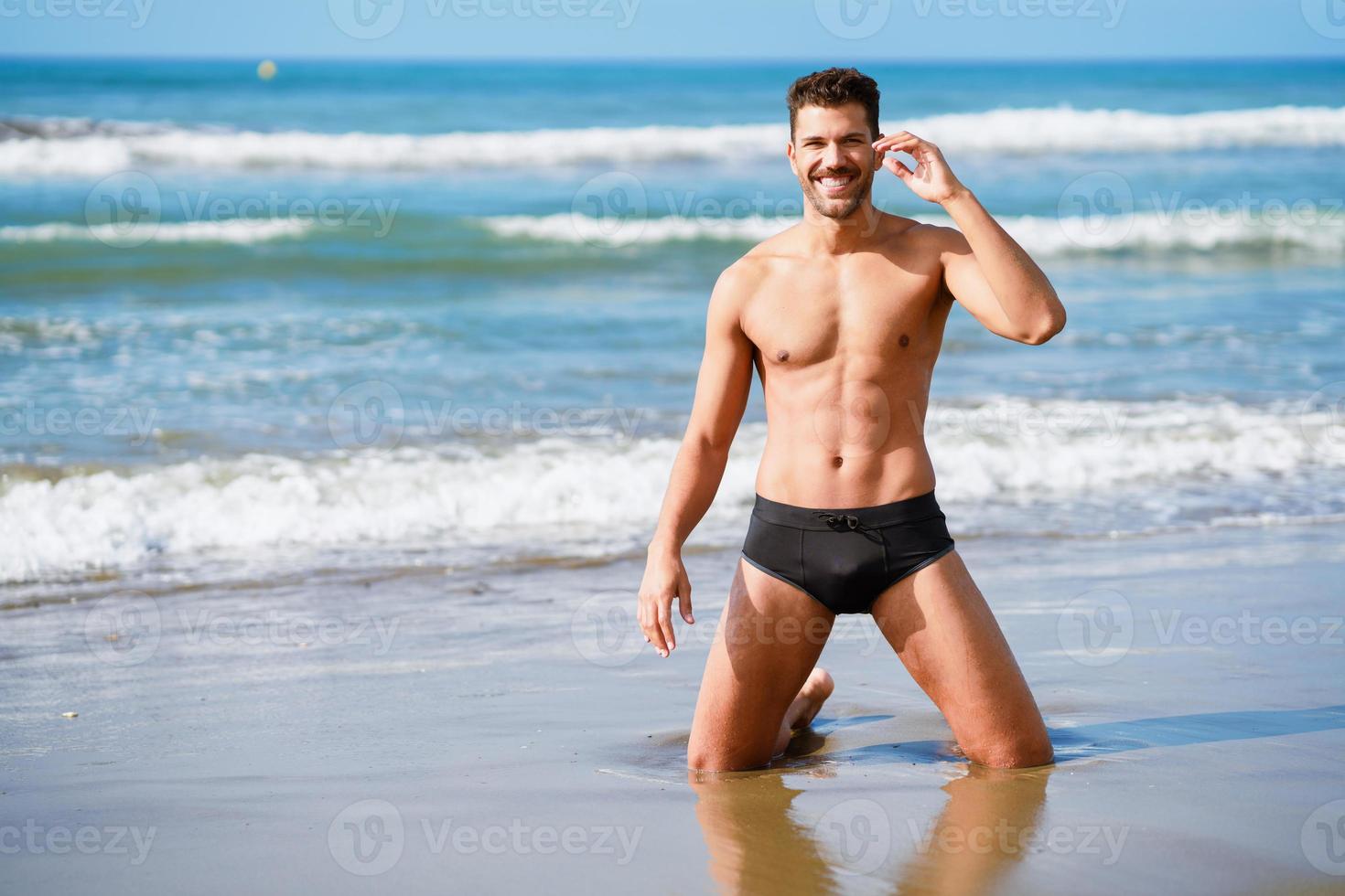 homem bonito de joelhos sorrindo na areia da praia foto