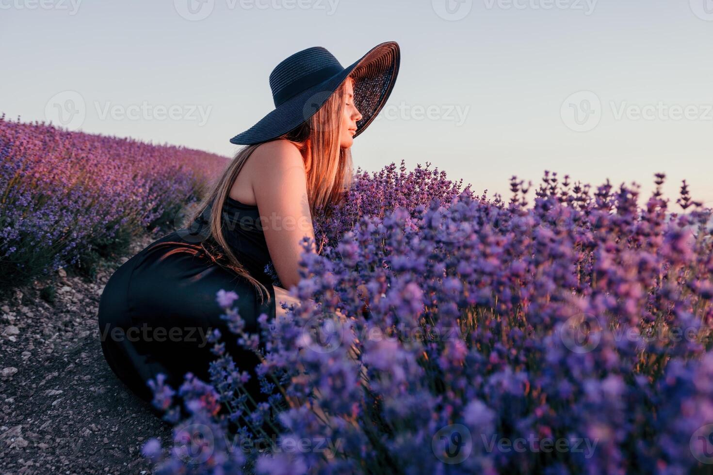 mulher lavanda campo. feliz despreocupado mulher dentro Preto vestir e chapéu com ampla borda cheirando uma florescendo lavanda em pôr do sol. perfeito para inspirado e caloroso conceitos dentro viagem e desejo de viajar. fechar acima foto