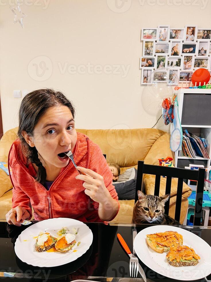 jovem mulher comendo torrada com salmão, abacate, caçado ovo e queijo enquanto sentado às a mesa às casa foto