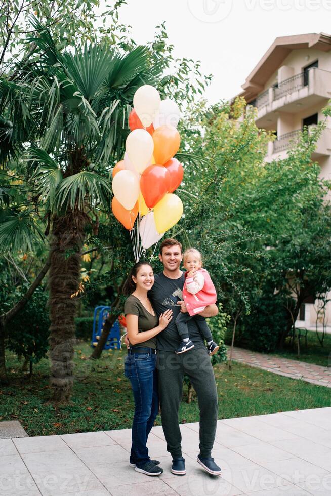 sorridente mãe abraçando Papai com uma pequeno menina dentro dele braços dentro a jardim perto colorida balões foto