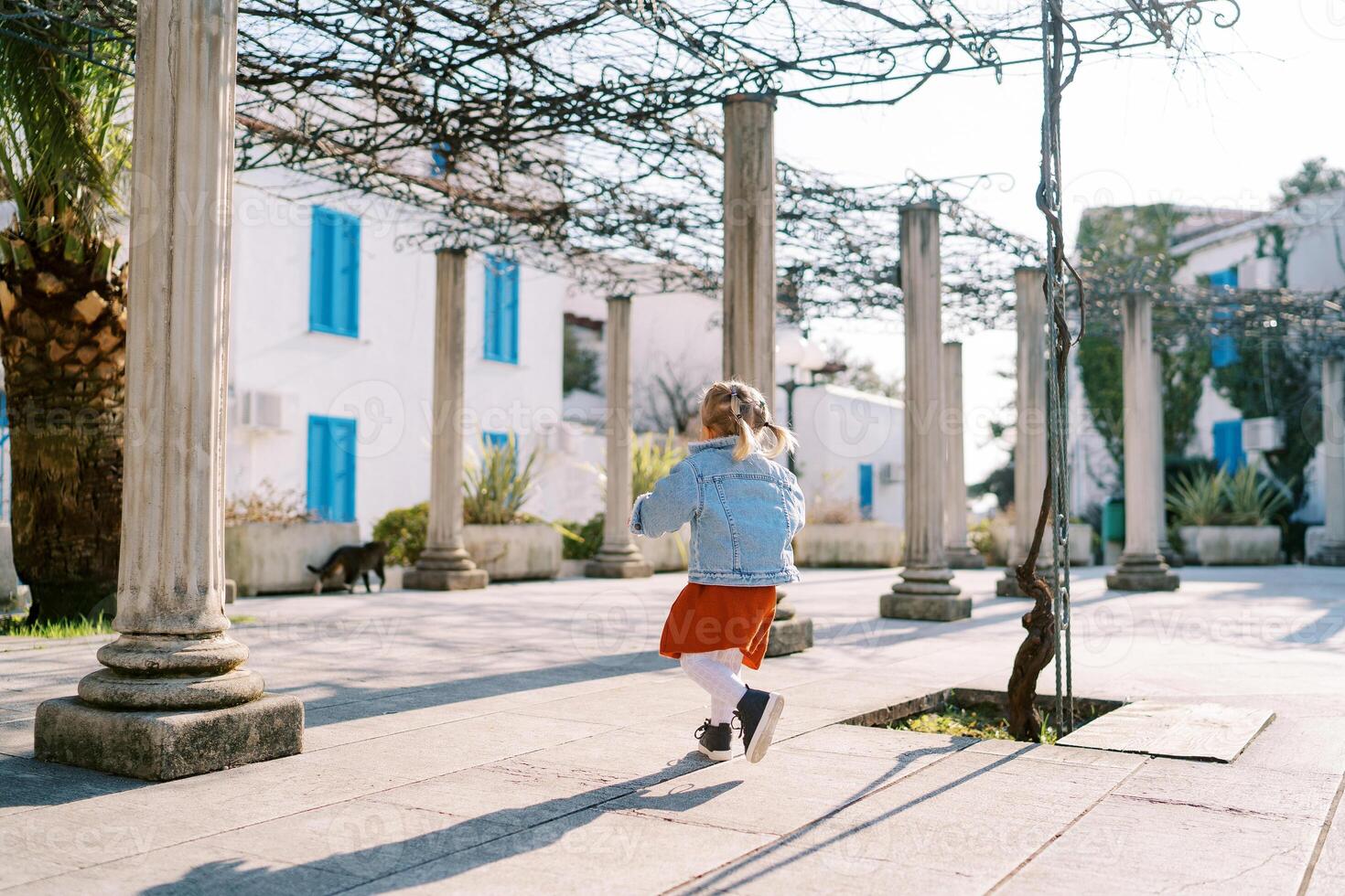 pequeno menina anda em entre pérgola colunas dentro uma jardim perto uma três andares construção foto