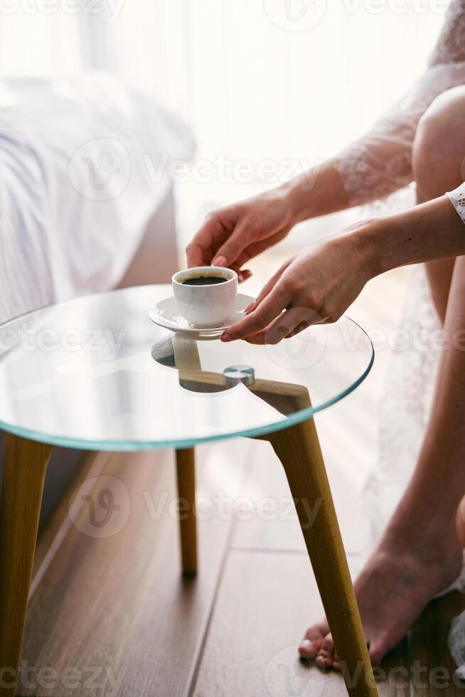 noiva leva uma copo do café a partir de uma vidro mesa enquanto sentado dentro a cadeirão. cortado. sem rosto foto