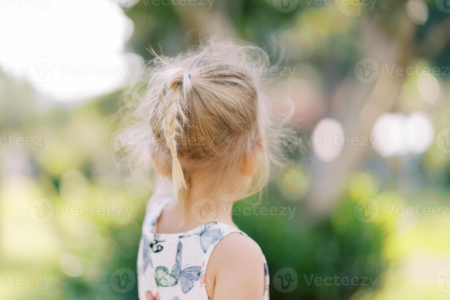 pequeno menina com fluindo cabelo carrinhos perto uma árvore e pontos para isto. costas Visão foto