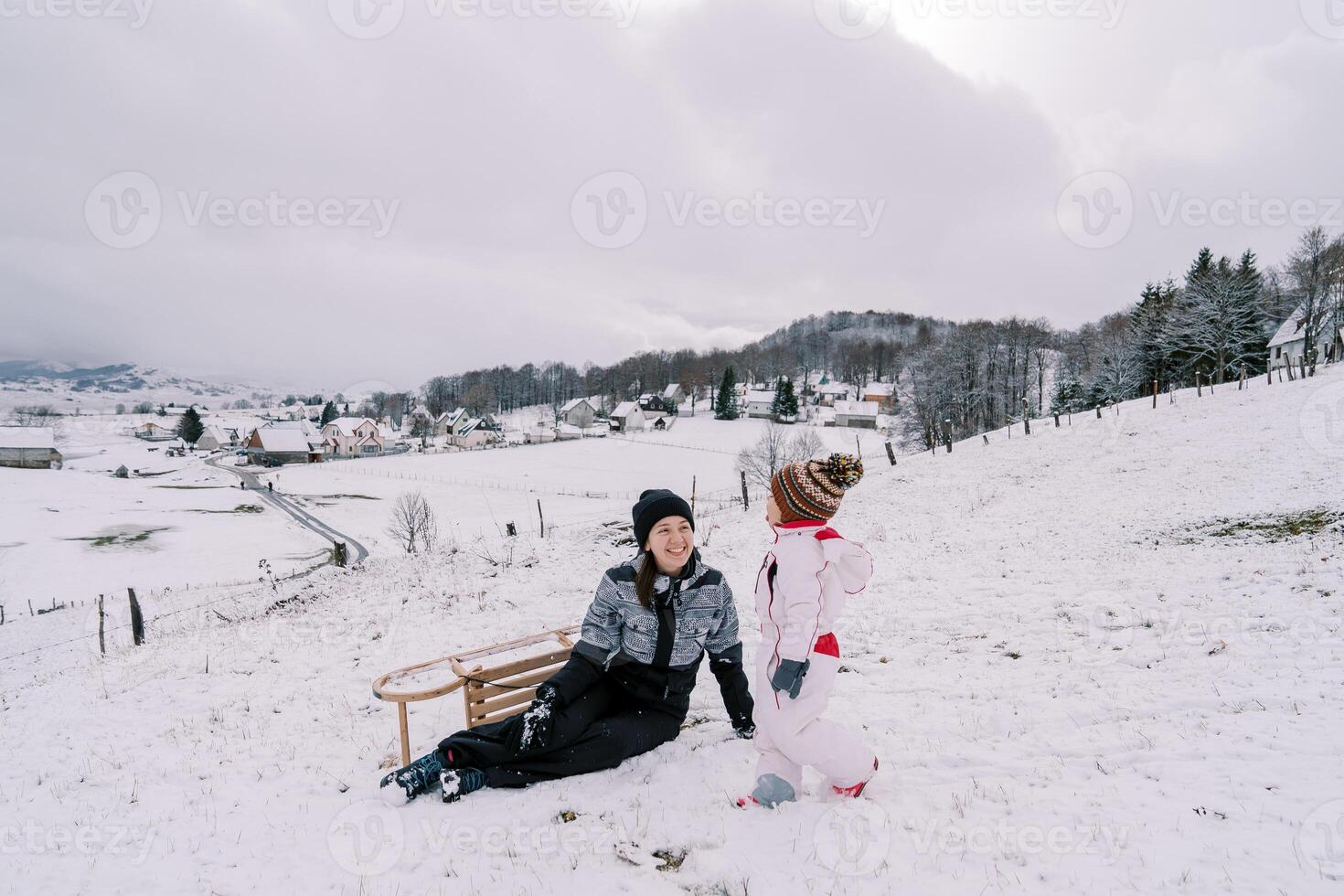 pequeno menina anda em ao longo uma Nevado Colina para dela sentado sorridente mãe com uma trenó virou em Está lado foto