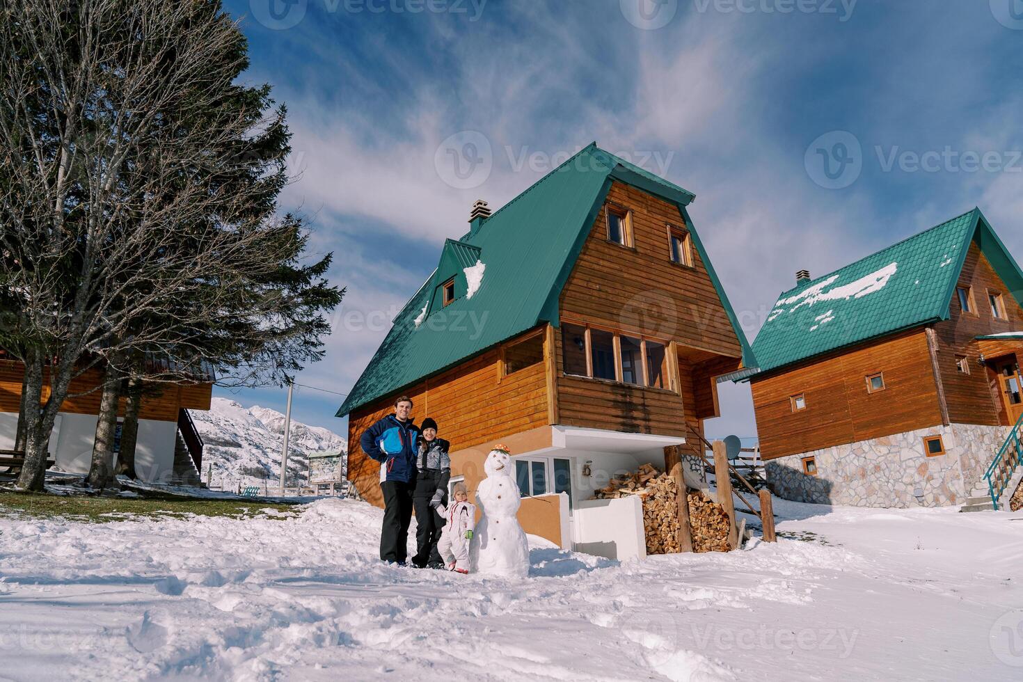 pai, mãe e pequeno menina ficar de pé perto uma boneco de neve Próximo para uma de madeira chalé foto