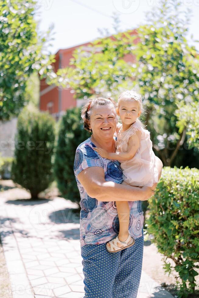 pequeno menina sentado dentro a braços do uma sorridente avó dentro a jardim foto