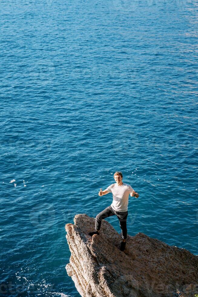 jovem sorridente homem em pé em uma pedra borda acima a mar com dele polegares acima foto