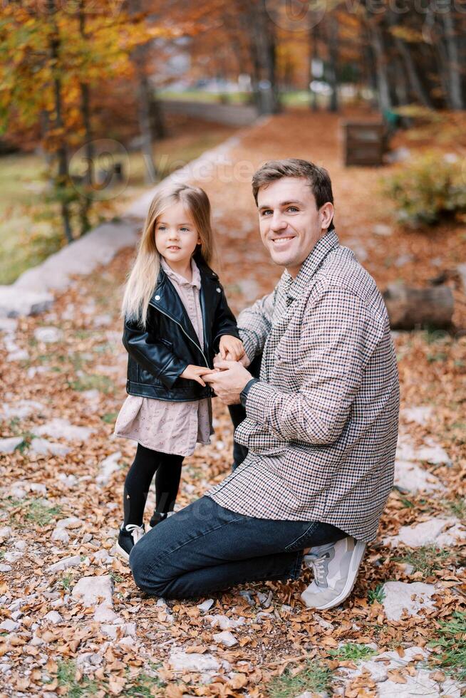 sorridente Papai segurando mãos com pequeno filha enquanto agachado dentro outono parque foto