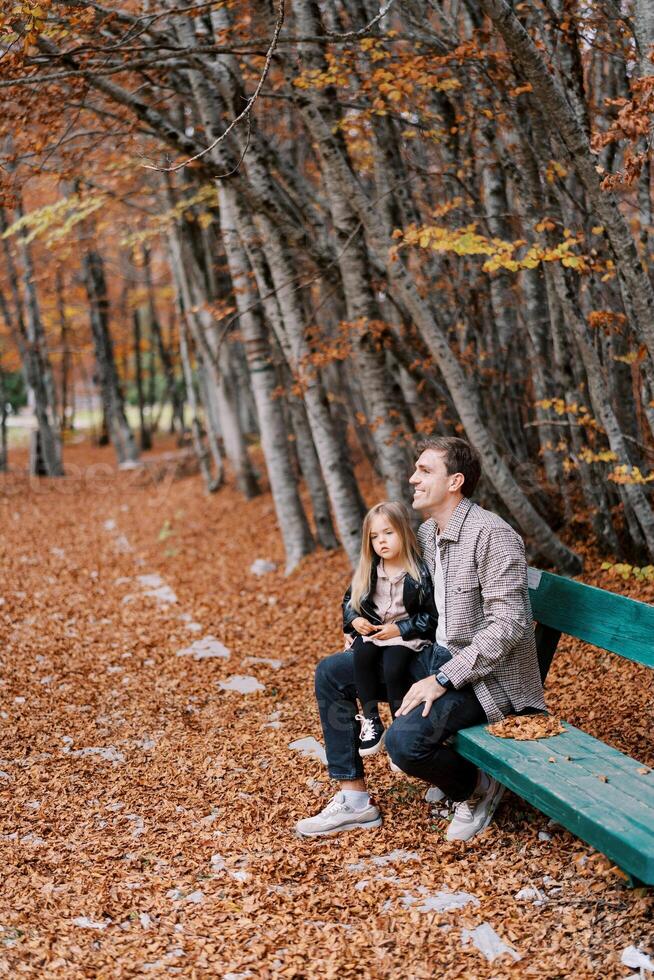 pequeno menina senta em a colo do uma sorridente Papai em uma Banco dentro a outono floresta foto