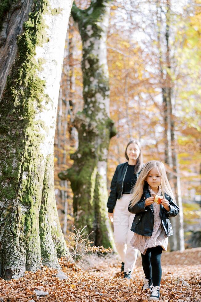 mãe segue uma pequeno menina roendo a maçã dentro a outono floresta foto