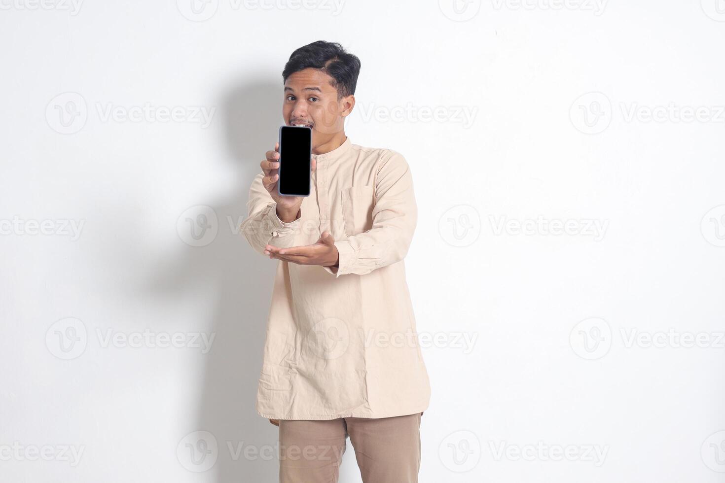 retrato do jovem animado ásia muçulmano homem dentro Koko camisa mostrando em branco tela Móvel telefone brincar enquanto apontando e apresentando produtos. social meios de comunicação conceito. isolado imagem em branco fundo foto