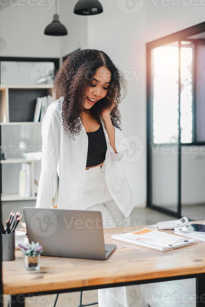 empresária dentro uma relaxado postura chats em a telefone, dela expressão 1 do prazer e noivado, dentro dela moderno escritório com uma computador portátil aberto antes dela. foto