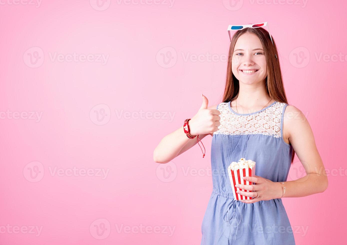 menina adolescente sorridente usando óculos 3D comendo pipoca isolada no fundo rosa aparecendo o polegar foto