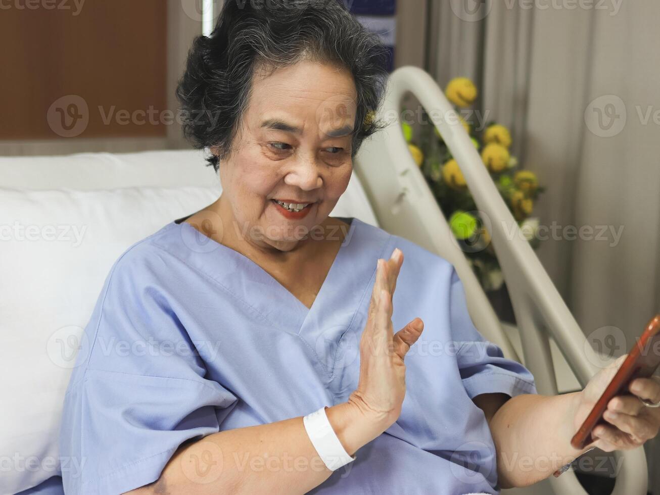 alegre ásia Senior fêmea paciente sentado dentro hospital cama, sorridente e olhando às Móvel telefone, vídeo chamando e acenando para dela família. idosos saúde conceito. foto
