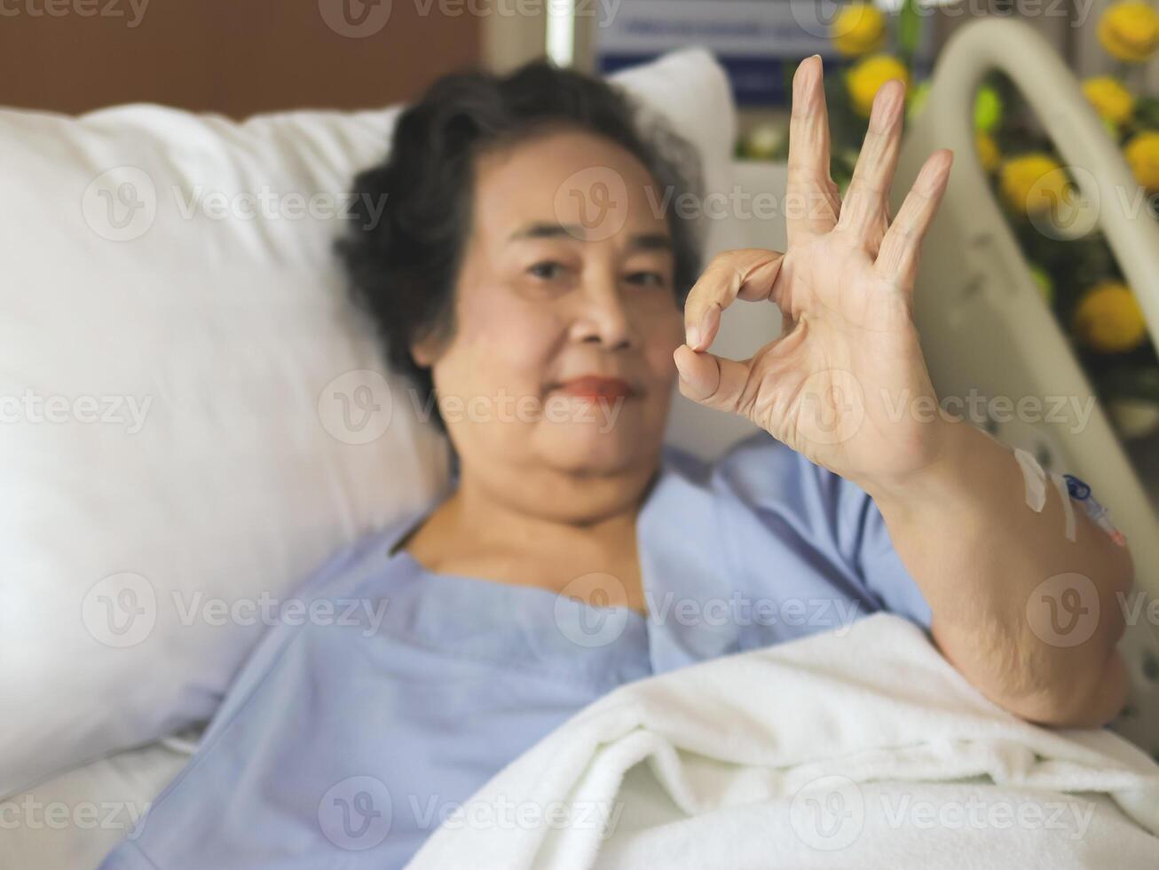 ásia Senior fêmea paciente deitado dentro hospital cama, sorridente e mostrando OK mão placa. idosos saúde conceito. seletivo foco em dedos. foto