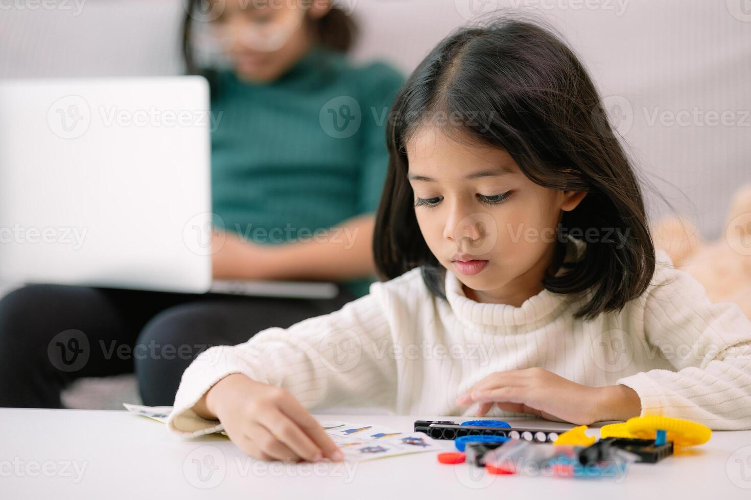 uma jovem menina é sentado às uma mesa com uma computador portátil e uma pilha do legos foto