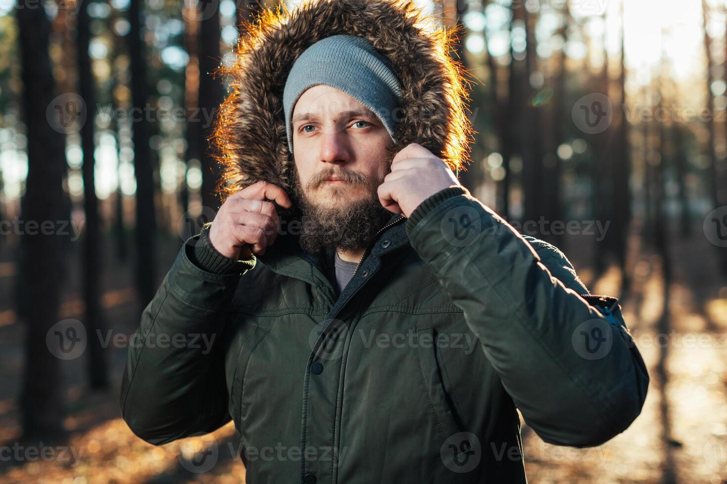 retrato, fechar-se do jovem elegantemente sério homem com uma barba vestido dentro verde inverno Jaqueta com uma de capuz e pele em dele cabeça carrinhos contra a fundo do pinho floresta. foto