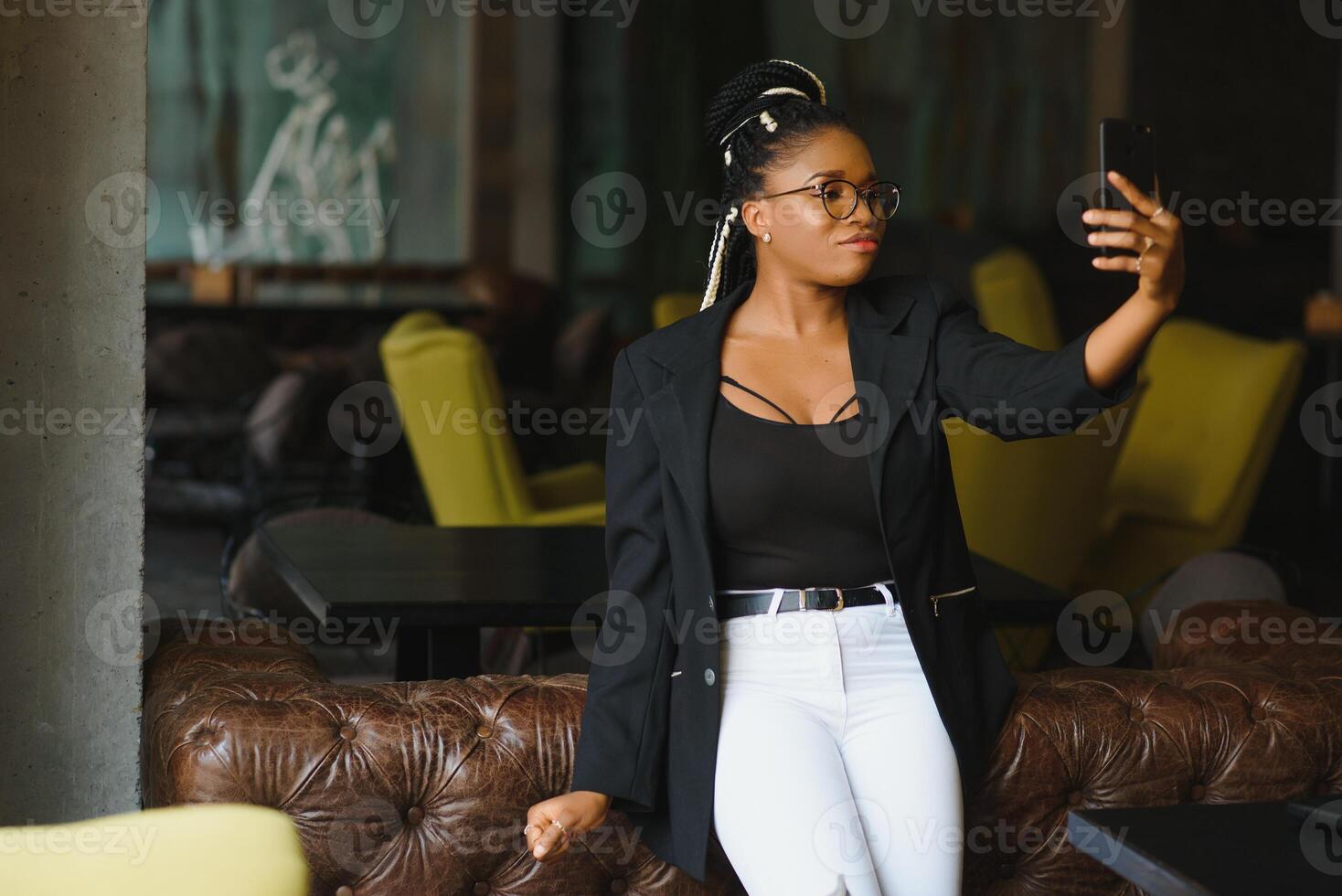 alegre Sombrio esfolado mulher dentro na moda vestem e óculos cafeteria posando para cenário e meios de comunicação conteúdo para blog, sorridente africano americano fêmea 20s fazer selfie usando Móvel telefone foto